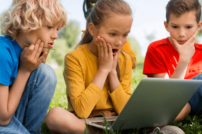 Drie kinderen kijken zittend in het gras naar een laptop. Ze schrikken van iets dat ze op sociale media zien. 