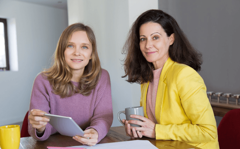 Twee vrouwen zitten aan tafel en kijken in de camera met een tablet en een kop thee en hebben een gesprek over de overgang