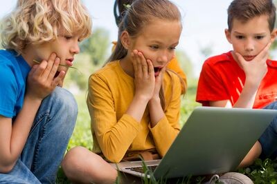 Drie kinderen kijken zittend in het gras naar een laptop. Ze schrikken van iets dat ze op sociale media zien. 