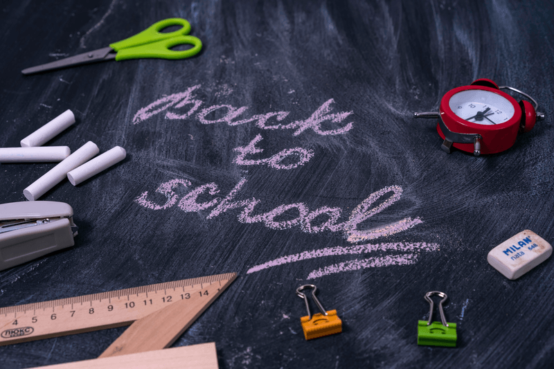 Abstracte weergave van een schoolbord waarop 'back to school' geschreven staat. Er liggen onder andere krijtjes, een schaar, een wekker en een gum op het schoolbord.